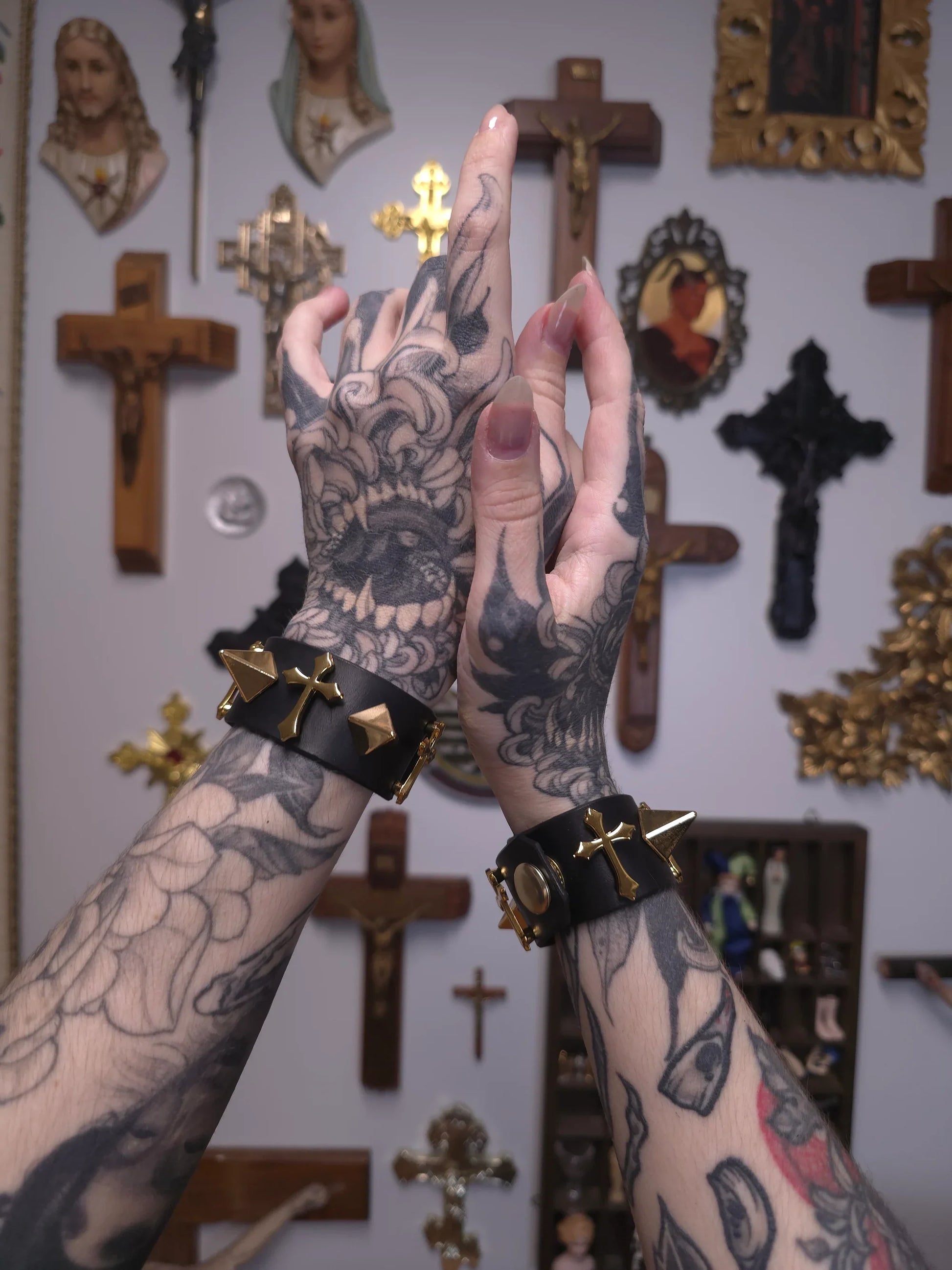 Tattooed hands with black leather bracelets in front of a wall with crosses and religious statues.