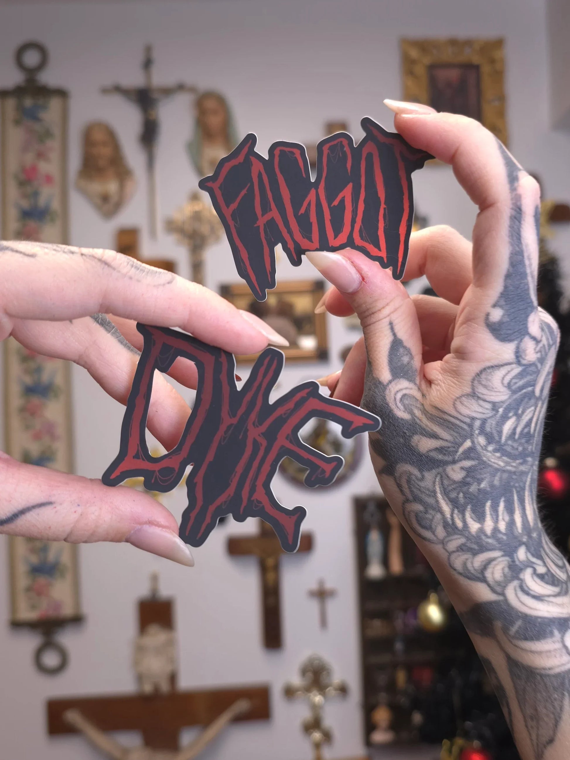 hands holding stickers with 'FAGGOT' 'DYKE' in metal logo fonts 