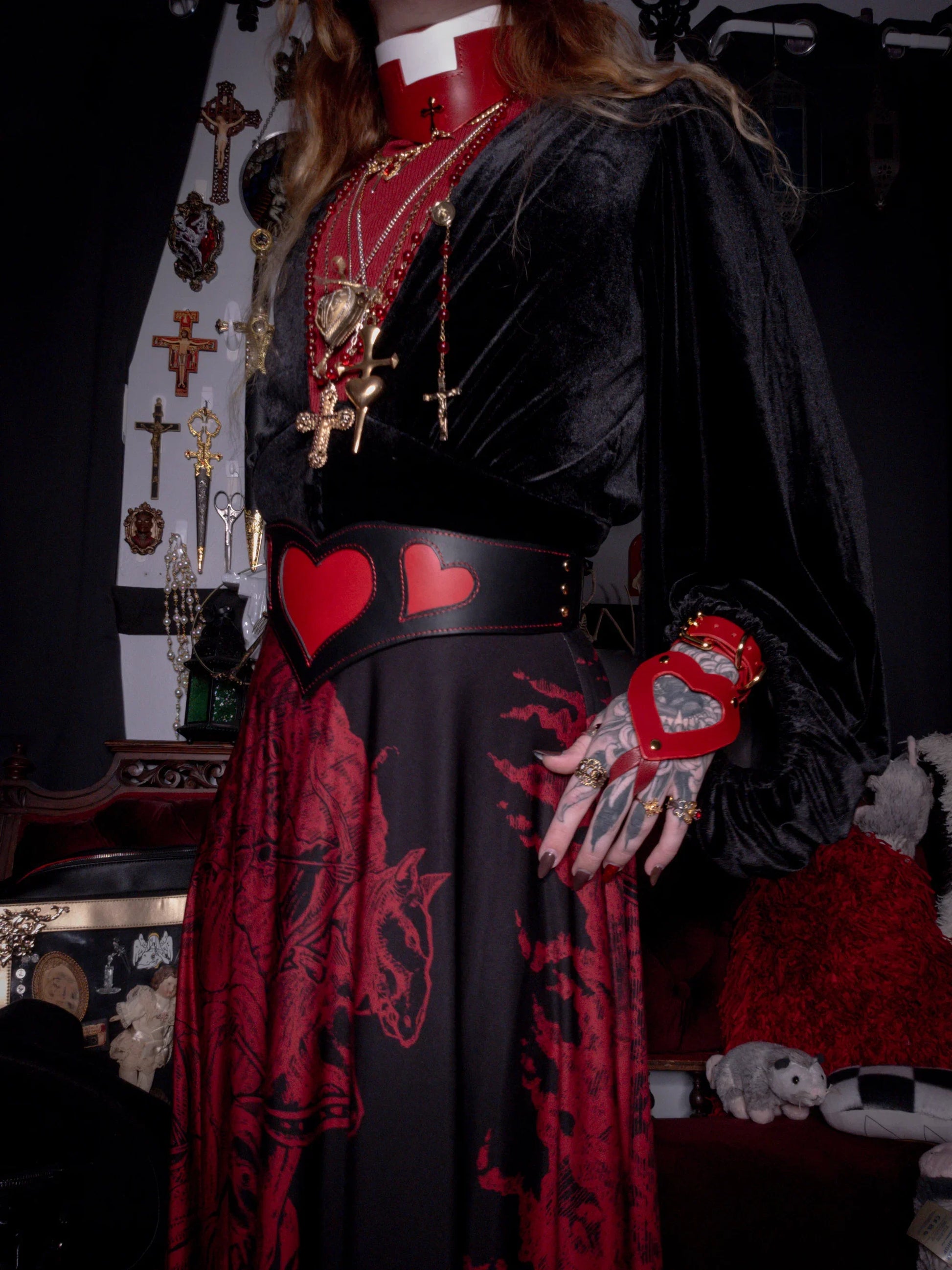 Person wearing a dark outfit with red accents and heart designs, standing in a dimly lit room.
