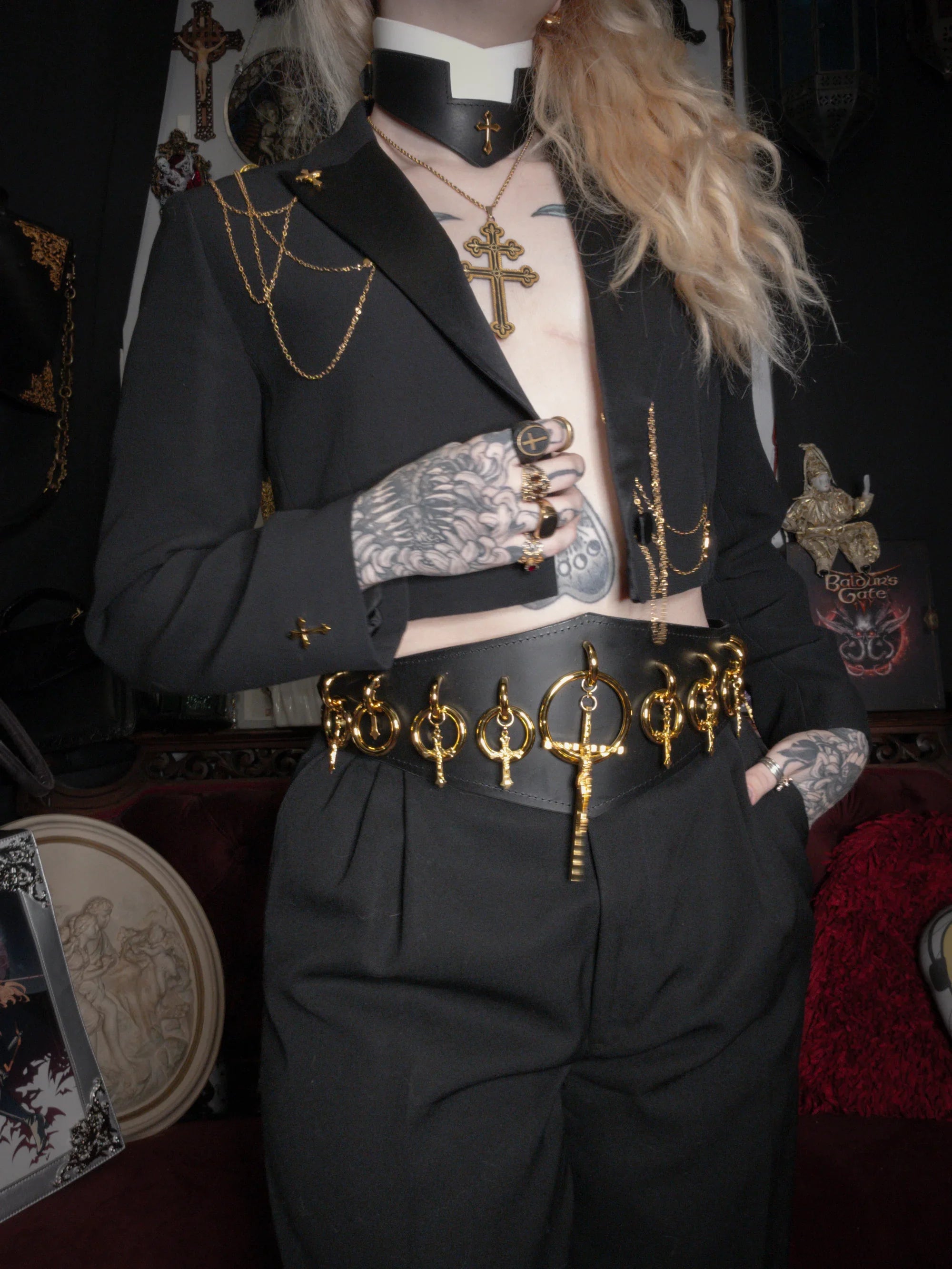Person wearing a black outfit with gold accents and tattoos, standing in a dark room.