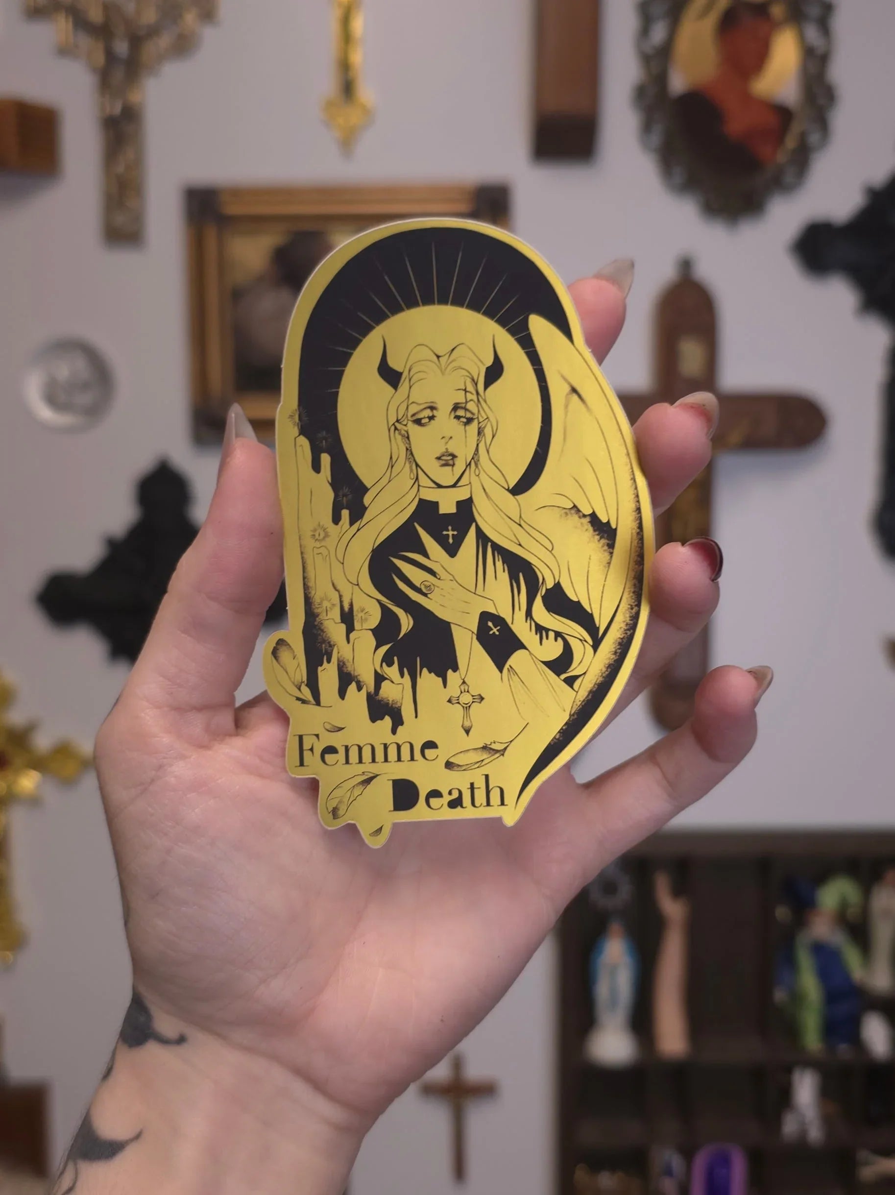 Hand holding a gold 'Femme Death' sticker with a black illustration against a blurred background.