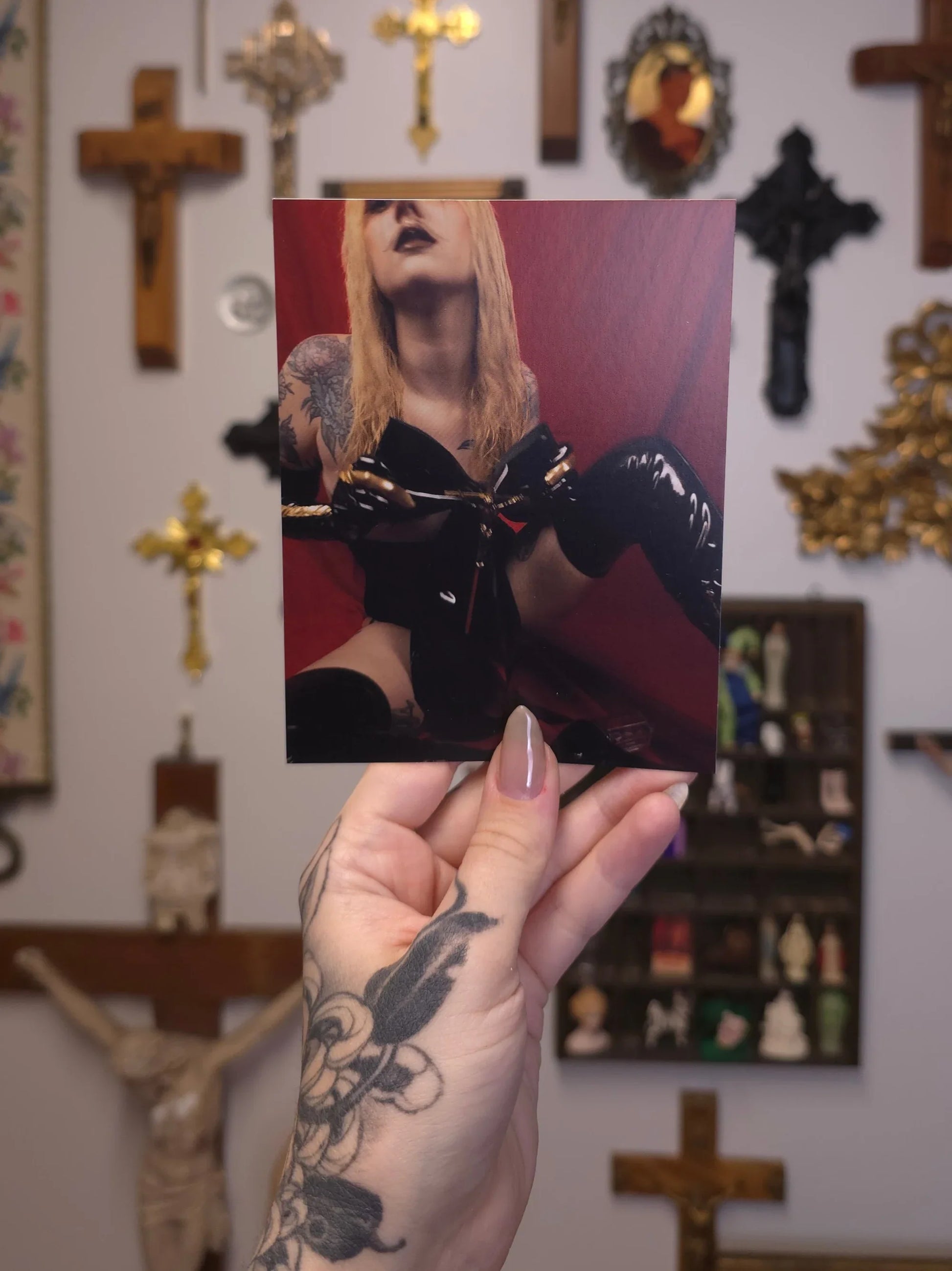 Hand holding a photo of a person in black leather outfit with tattoos, against a background of religious statues and crosses.