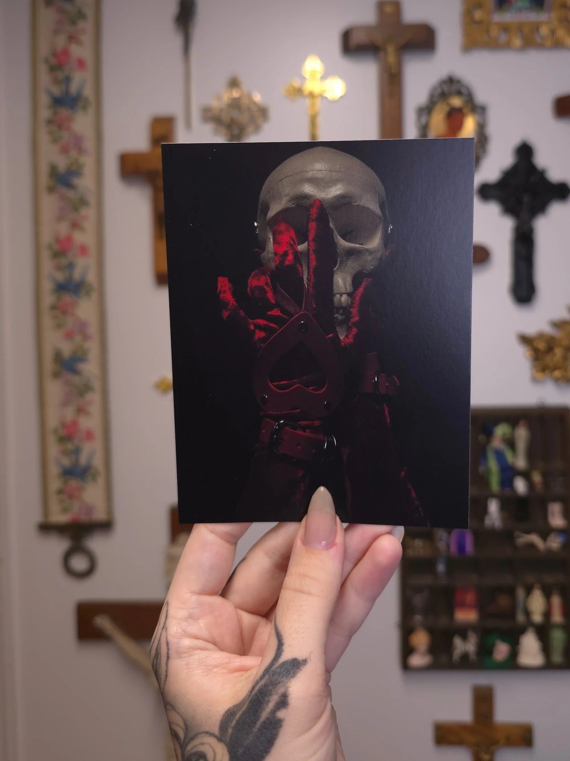 Person holding a card with a red and black design featuring a skull against a dark background, with a blurred background of religious items.