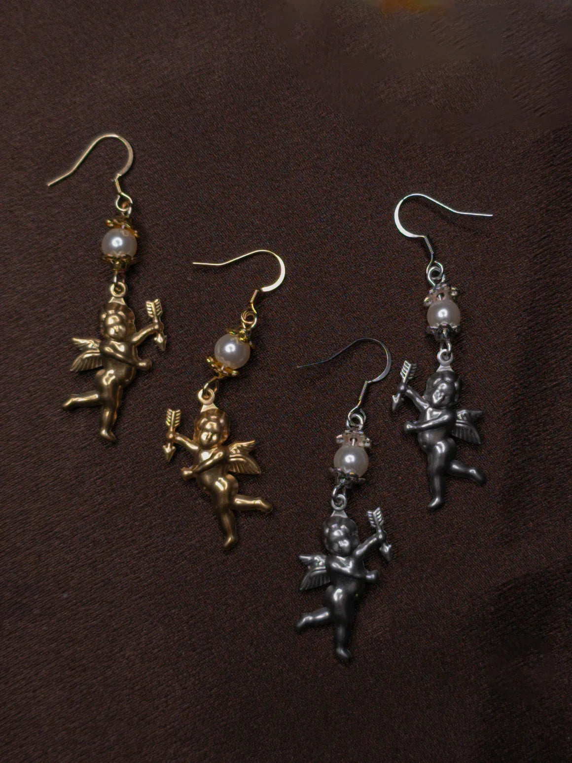 Two pairs of angel-shaped earrings in gold and silver on a dark brown background.