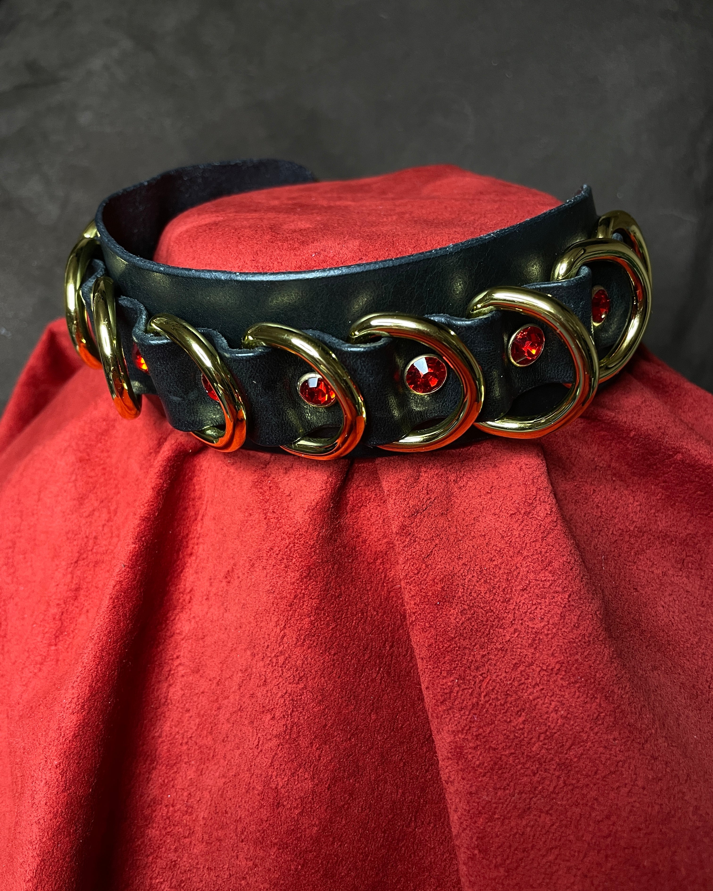 Black leather collar with gold rings and red stones on a red cushion