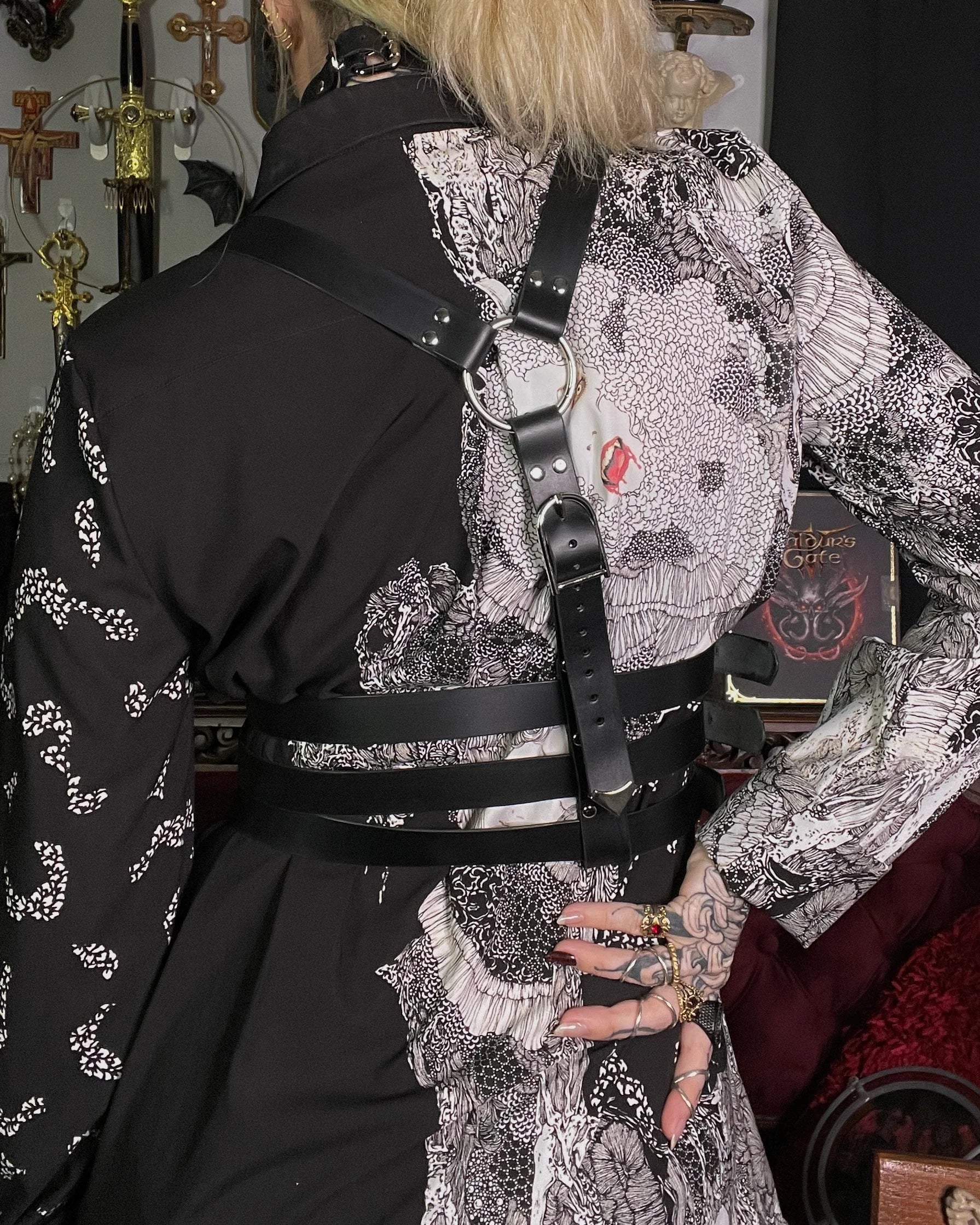 Person wearing a black and white patterned outfit with a harness, sitting in a room with decorative items.