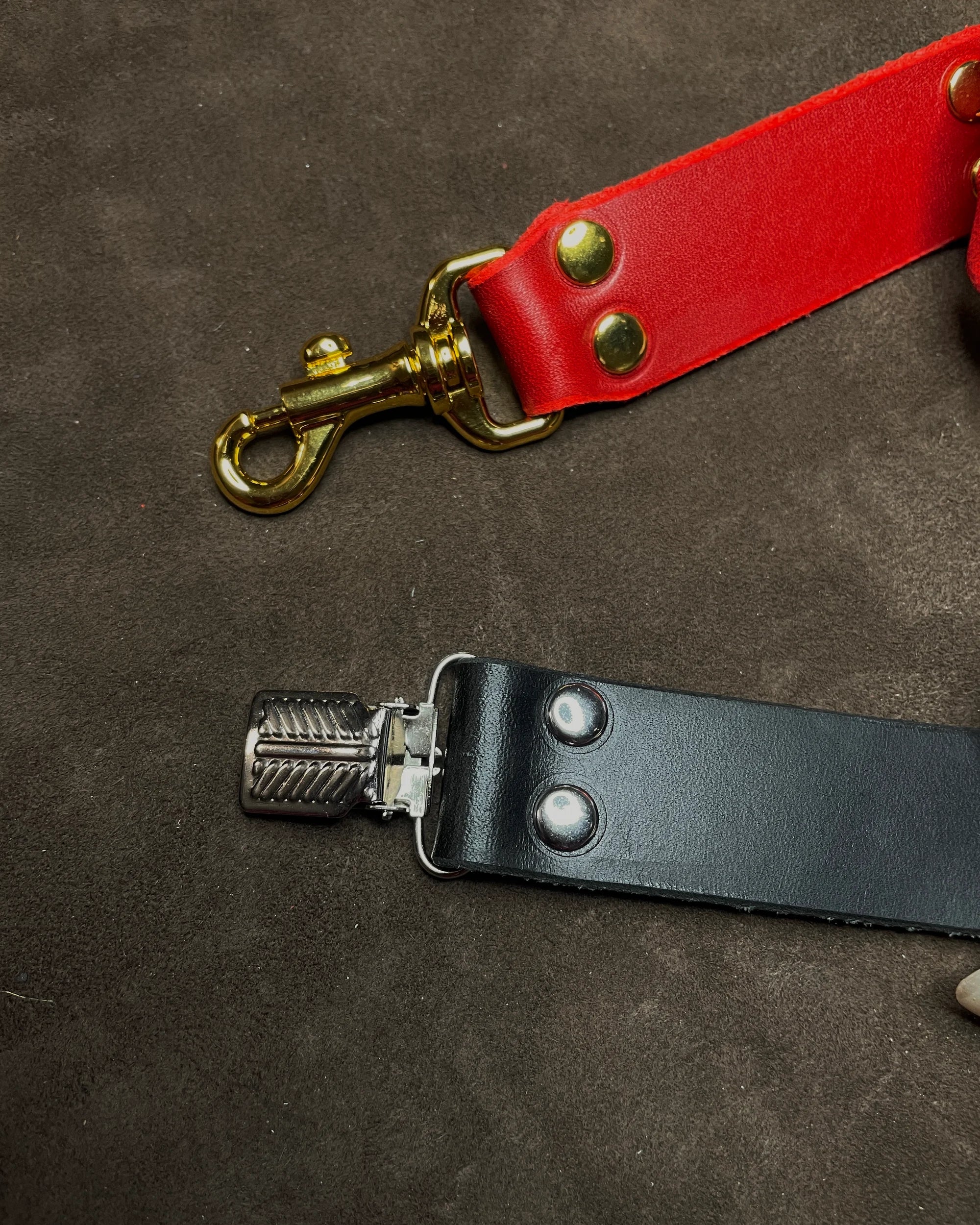 Close-up of a red leather strap with gold hardware and a black leather strap with metal clip on a brown background.