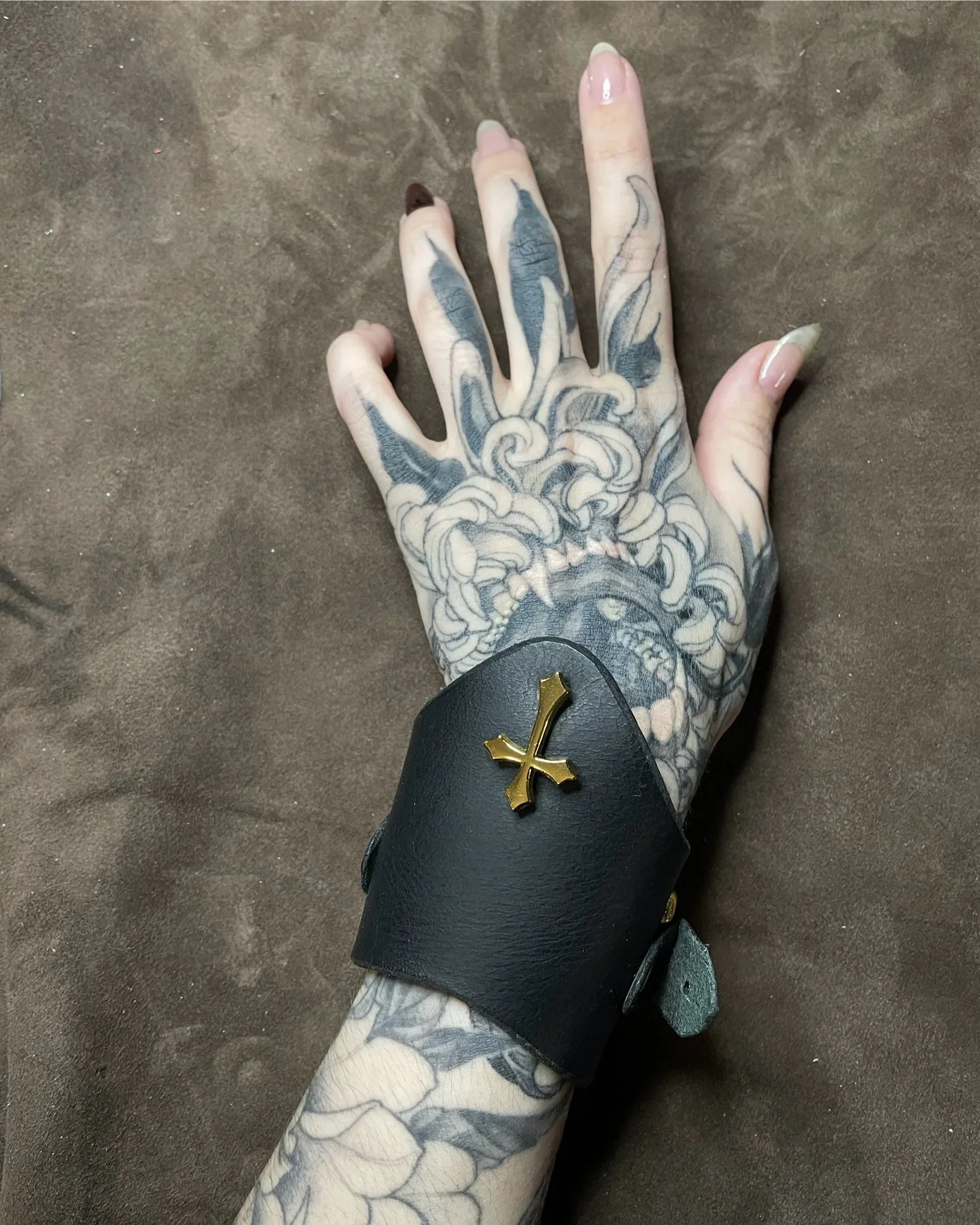 Tattooed hand wearing a black leather cuff with a gold cross on a brown background