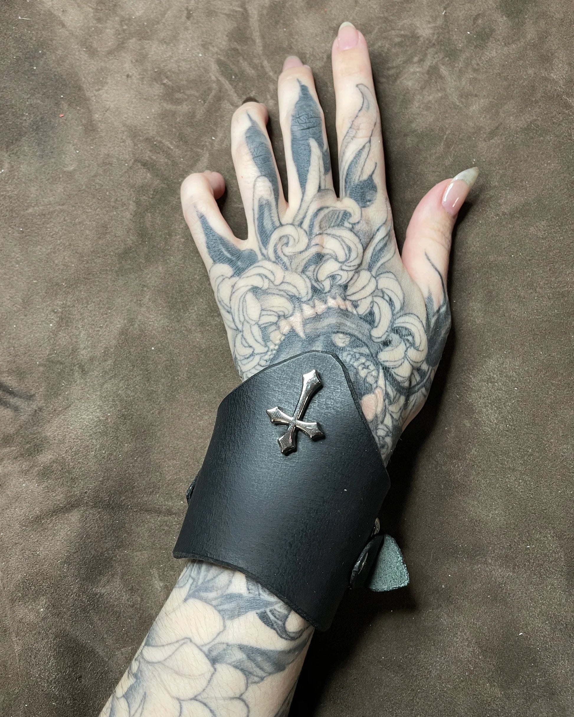 Tattooed hand wearing a black leather cuff with a silver cross on a brown background