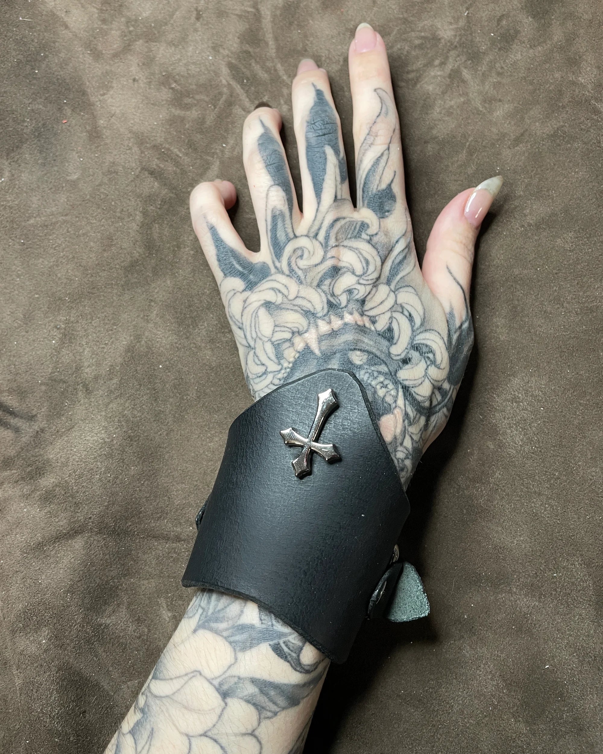 Tattooed hand wearing a black leather cuff with a silver cross on a brown background