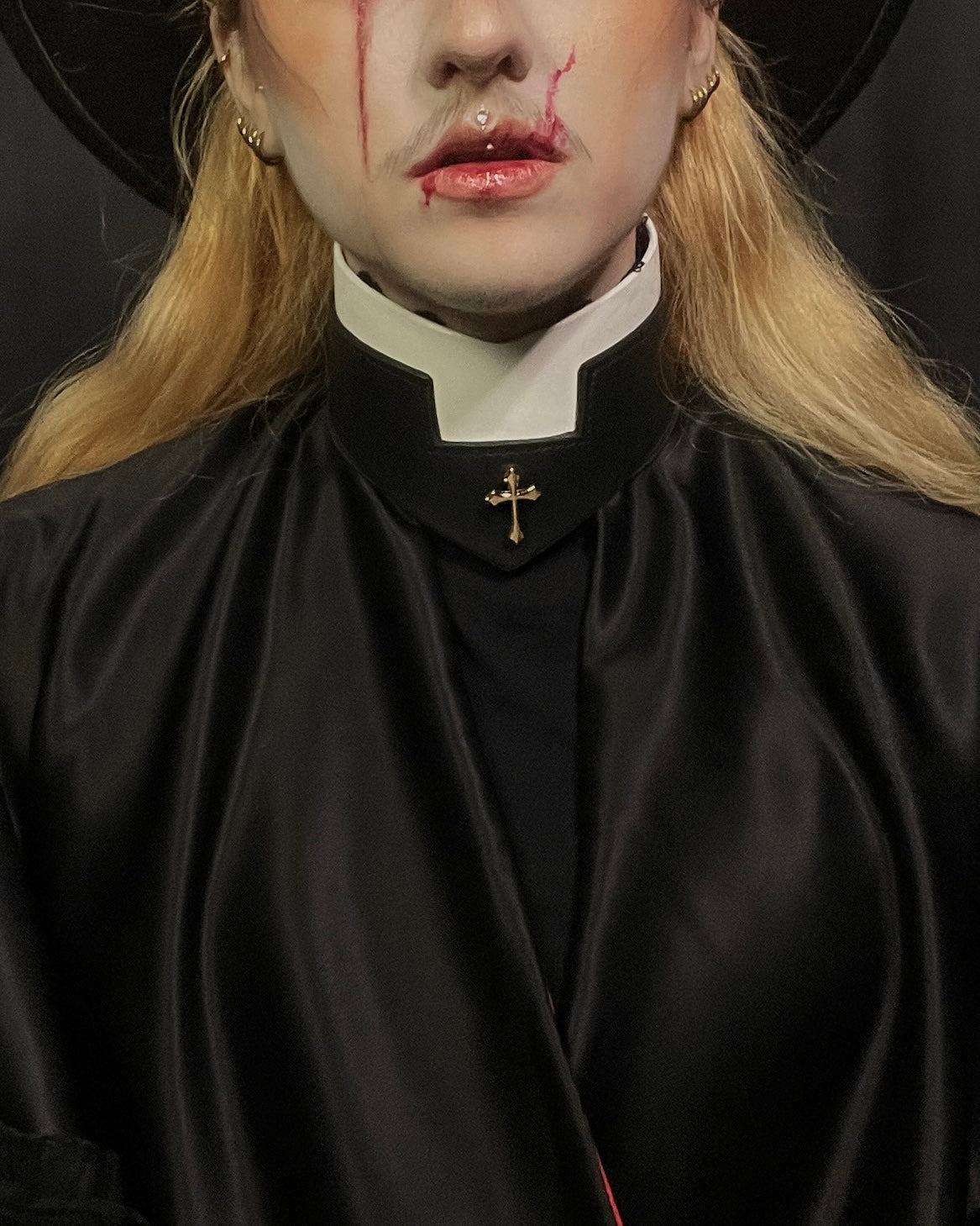 Person wearing a black robe with a cross emblem, resembling a religious figure, on a dark background.