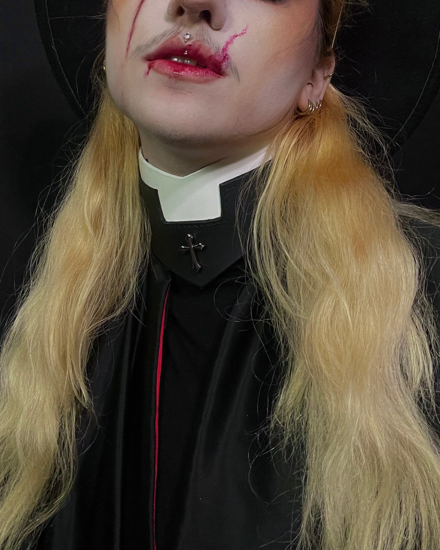 Person with blonde hair wearing a black outfit with a white collar against a dark background.