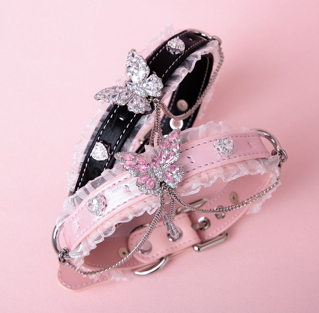 two black and pink leather collars featuring white ruffled lace trim, intricate crystal butterfly and heart charms, silver D rings and chains, displayed on a pink background.