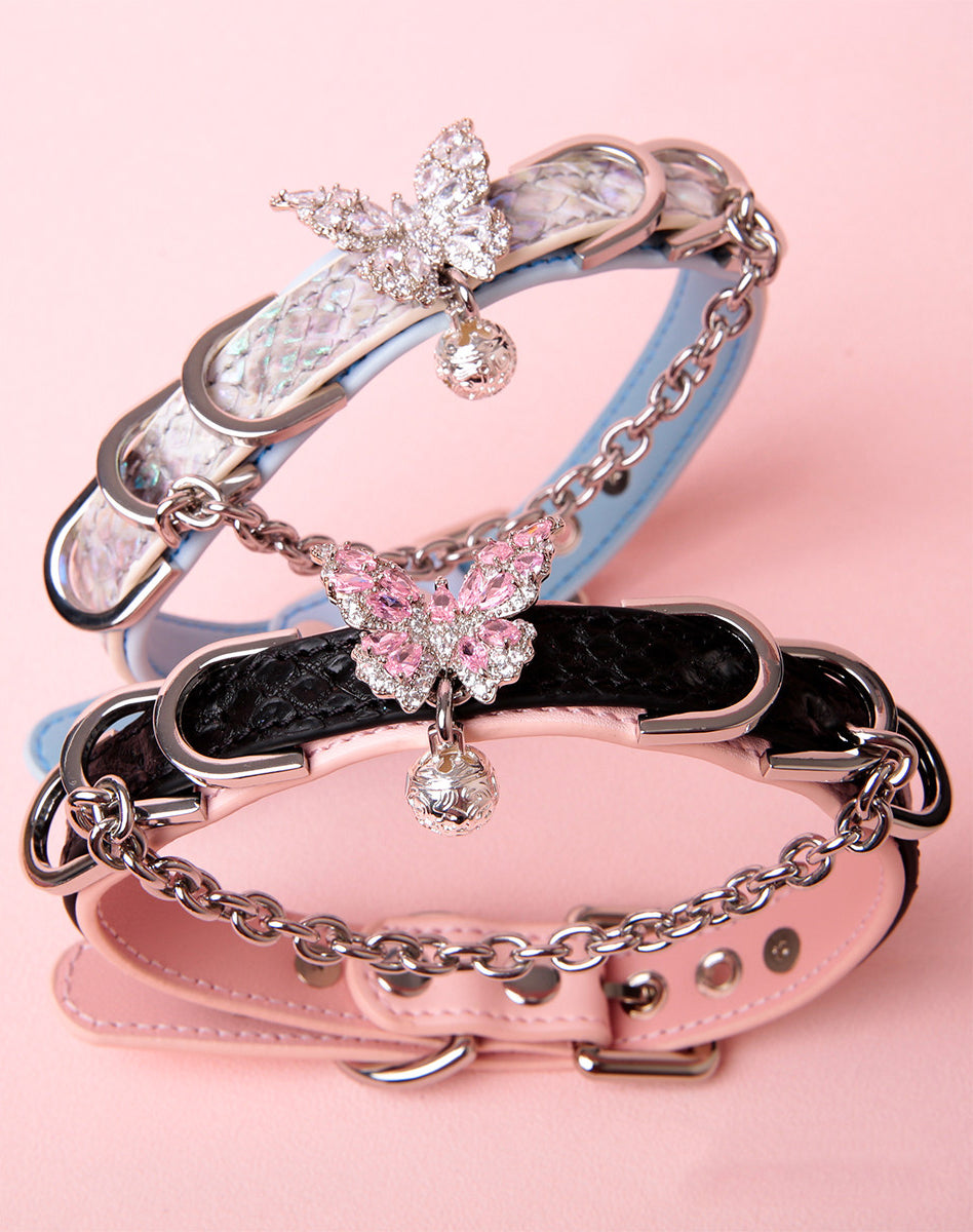 two black and blue leather collars featuring silver diamante butterfly pendants, chains, D rings and bell charms, with adjustable buckle fastening at the back, displayed on a pink background.