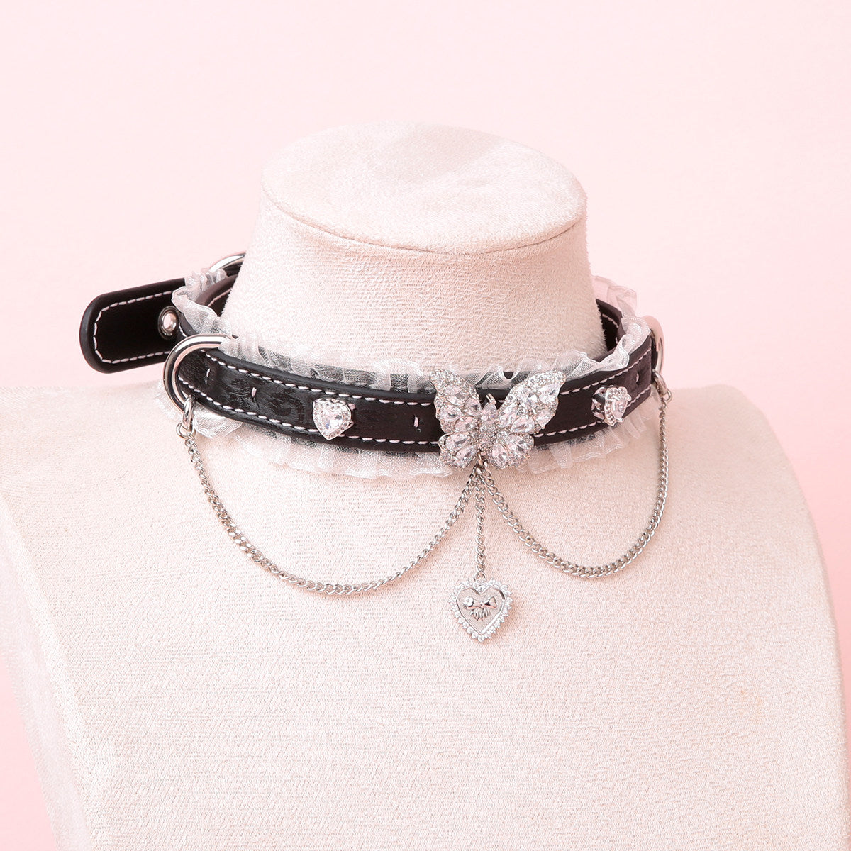 black leather collar featuring white ruffled lace trim and intricate silver butterfly pendant, D rings, chains and crystal heart charms, displayed on a white mannequin against a pink background.