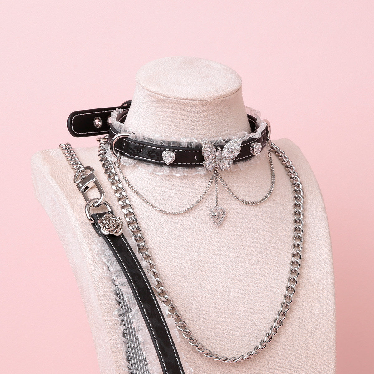 black leather collar and leash set featuring white ruffled lace trim and intricate silver butterfly and crystal heart charms, with a detachable chain leash and leather hand strap, displayed on a white mannequin against a pink background.