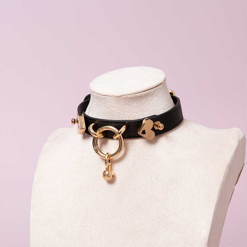 Black leather collar featuring a central gold tone O ring and detachable bell charm. Displayed on a mannequin against a pink background.
