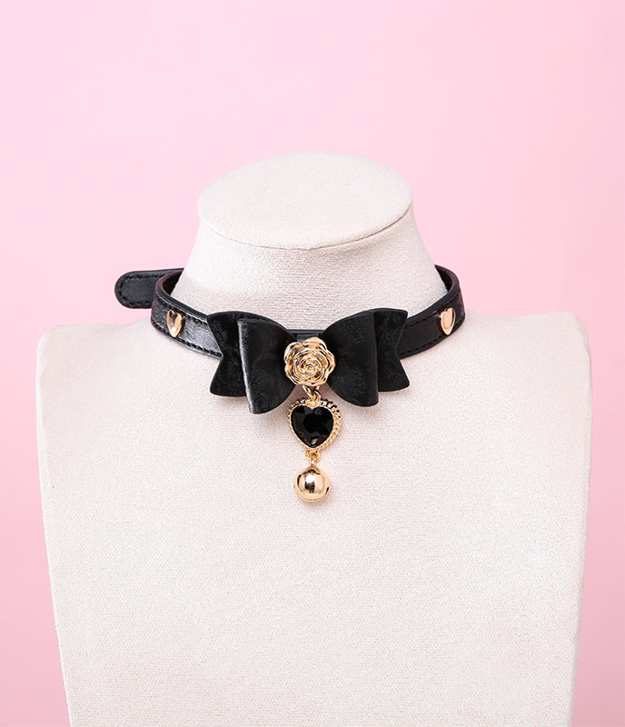 black and gold collar featuring a ribbon bow shaped centre, gold rose and heart studs with a black and gold crystal heart and bell charm, displayed on a white mannequin against a pink background.