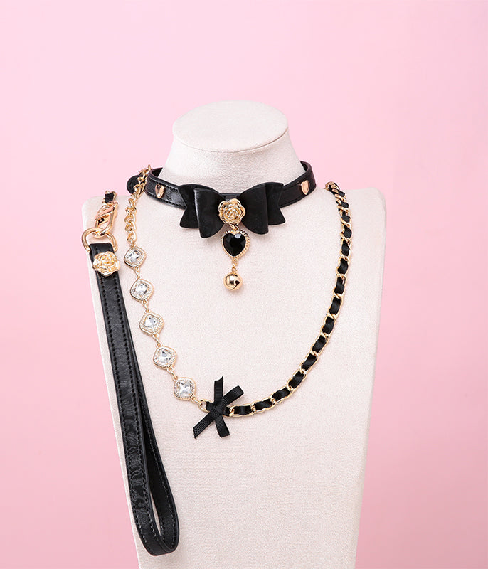 black and gold collar and leash set featuring ribbon bow shaped centre, gold rose and heart studs, black and gold crystal heart and bell charm with a detachable chain and crystal leash and leather hand strap, displayed on a white mannequin against a pink background.
