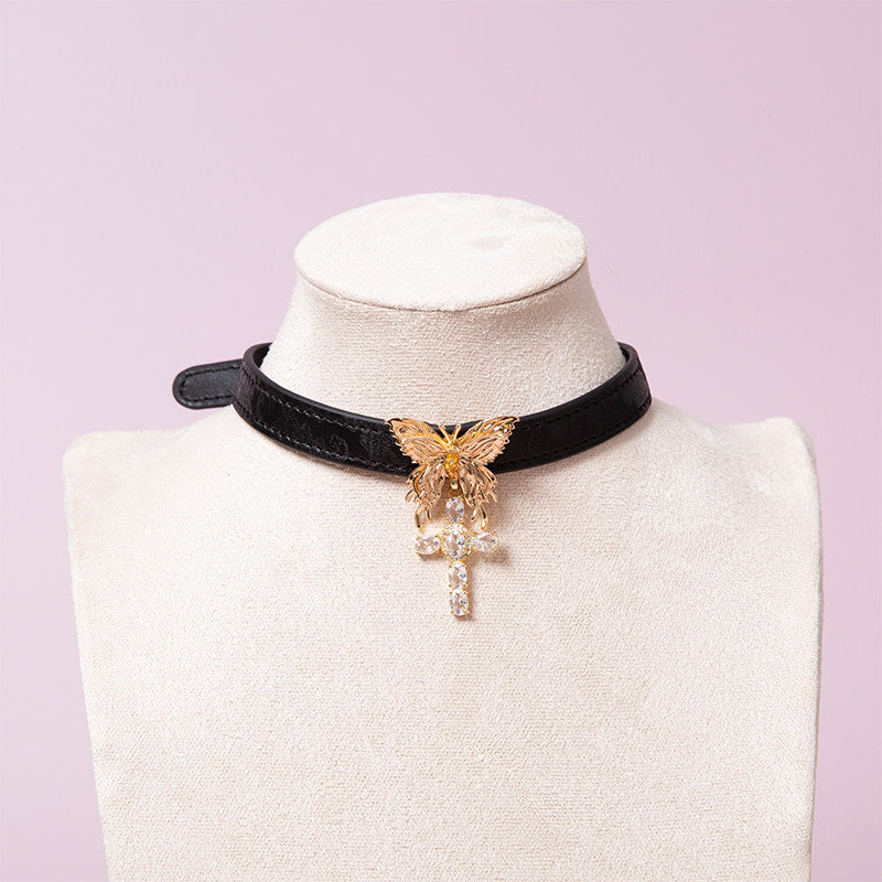 black and gold collar featuring a gold tone butterfly pendant and gold and diamante cross charm, displayed on a white mannequin against a pink background.