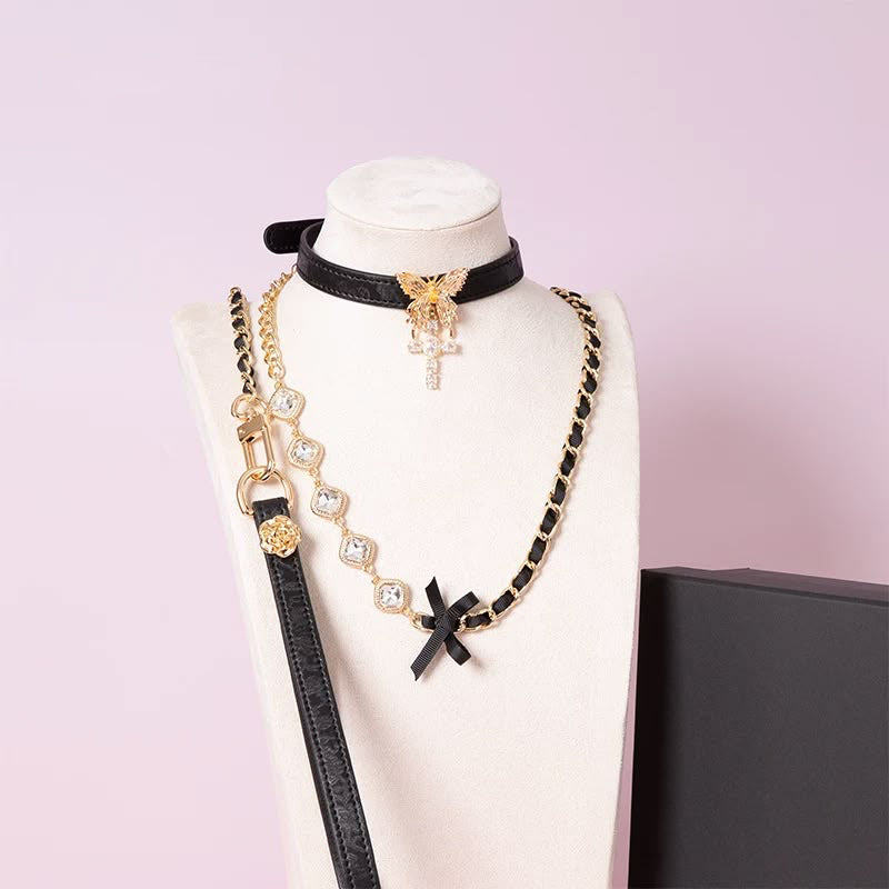 black and gold collar and leash set featuring a gold tone butterfly pendant, gold and diamante cross charm, detachable diamante and chain leash with black leather hand strap, displayed on a white mannequin against a pink background.