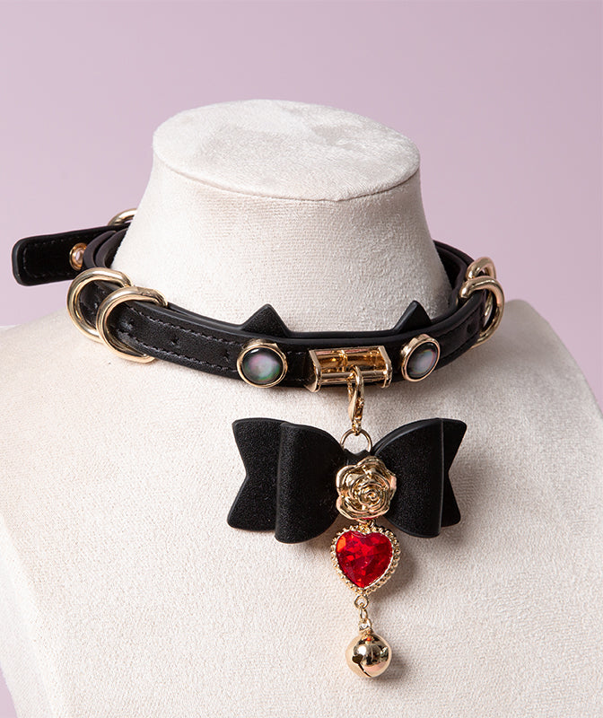 Black and gold cat girl collar featuring a unique cat ear shaped choker, gold tone hardware and detachable black and gold bow pendant with red crystal heart charm. Displayed on a mannequin against a pink background.