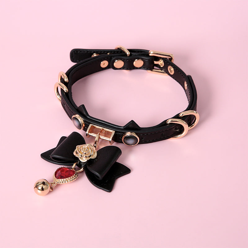 Black and gold cat girl collar featuring a unique cat ear shaped choker, gold tone hardware and detachable black and gold bow pendant with red crystal heart charm. Displayed on a pink background.