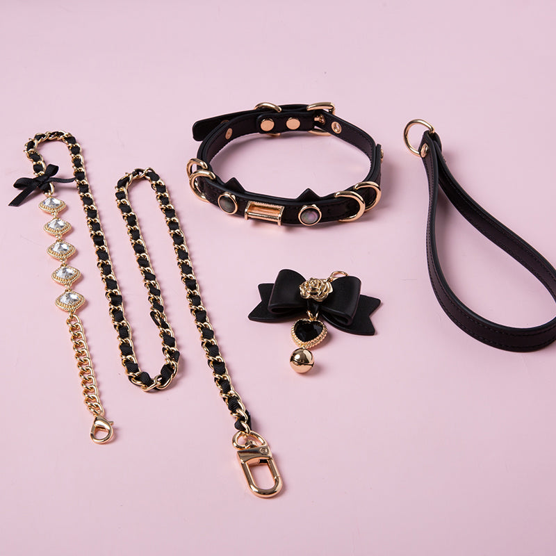 Black and gold cat girl collar and leash set featuring a unique cat ear shaped choker, detachable black and gold bow, heart and bell pendant, detachable crystal and chain leash and black leather hand strap. Displayed on a pink background.