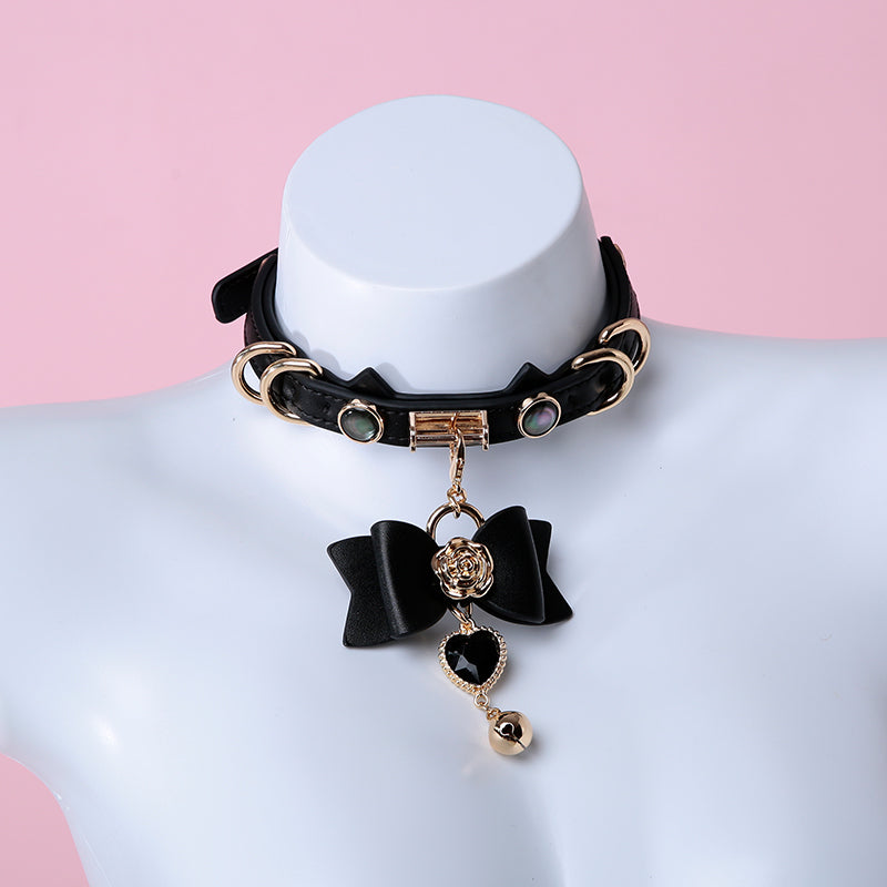 Black and gold cat girl collar featuring unique cat ear shaped choker strap, gold tone hardware and detachable black and gold bow, heart and bell pendant. Displayed on a mannequin against a pink background.