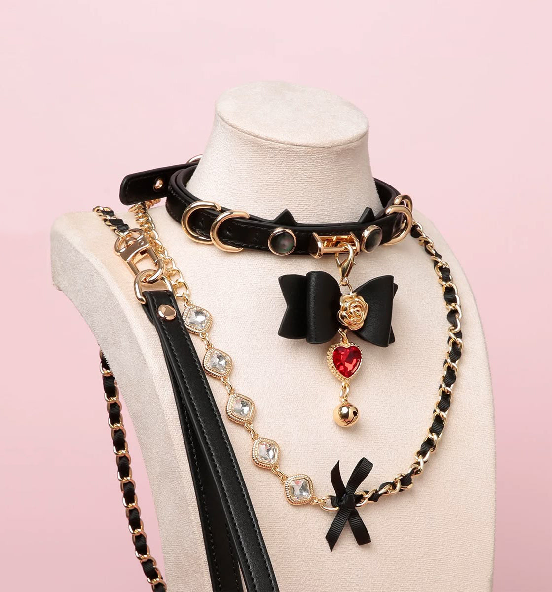 Black and gold cat girl collar and leash set featuring unique cat ear shaped choker, gold tone hardware, detachable bow and heart charm, crystal and chain leash and black leather hand strap. Displayed on a mannequin against a pink background.