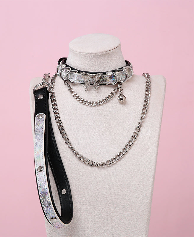 black and holographic silver collar and leash set featuring an ornate silver butterfly pendant, silver bell, D rings and detachable black and silver chain leash and leather hand strap, displayed on a white mannequin bust against a pink background.