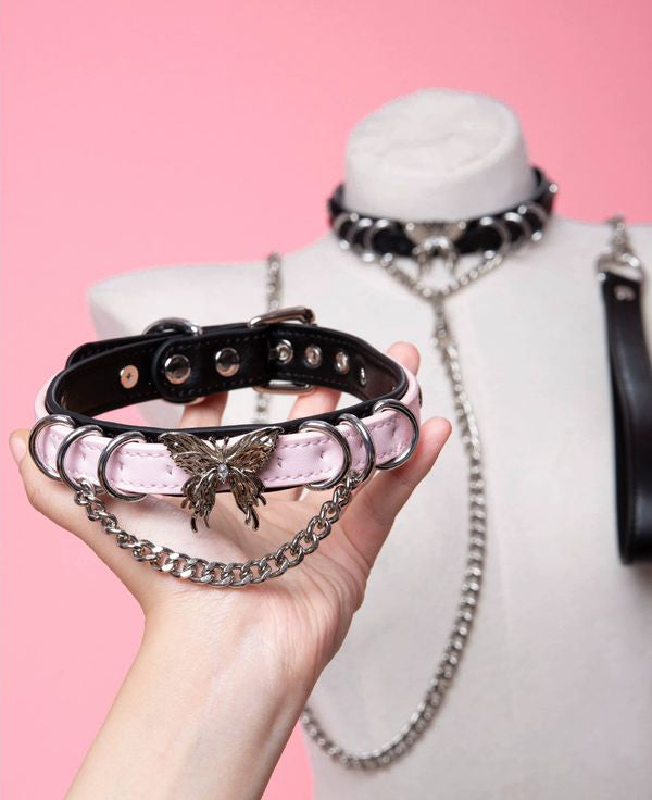 black and pink leather collar featuring an ornate silver butterfly pendant, silver bell, D rings and chain, held by a hand in front of a white mannequin against a pink background.