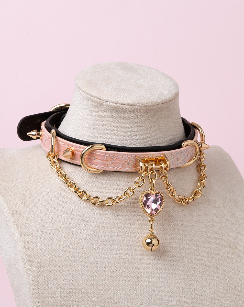 black, pink and gold collar featuring a pink and gold crystal heart and bell charm, gold chains and spike studs, displayed on a white mannequin bust against a pink background/
