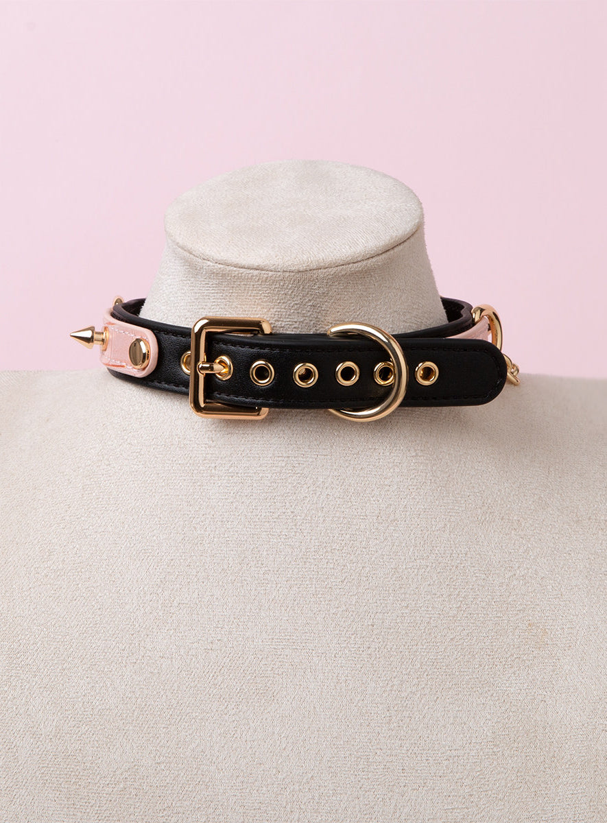 rear view of a black, pink and gold crystal heart collar, showing the adjustable gold tone buckle and eyelet holes, hardware and spike studs, displayed on a white mannequin bust against a pink background.