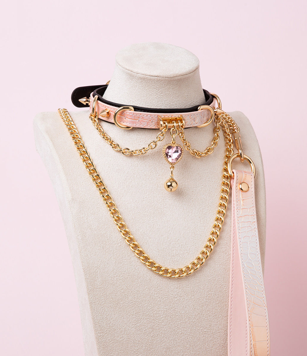 black, pink and gold collar and leash set featuring a pink and gold crystal heart and bell charm, gold chain leash and pink leather hand strap, displayed on a white mannequin bust against a pink background/