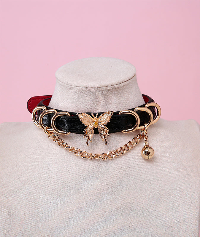 black, red and gold collar featuring an ornate gold butterfly pendant, gold bell, D rings and chain, displayed on a white mannequin bust against a pink background.