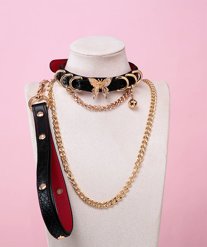 black, red and gold collar and leash set featuring an ornate gold butterfly pendant, gold bell, D rings and detachable black, red and gold chain leash and leather hand strap, displayed on a white mannequin bust against a pink background.