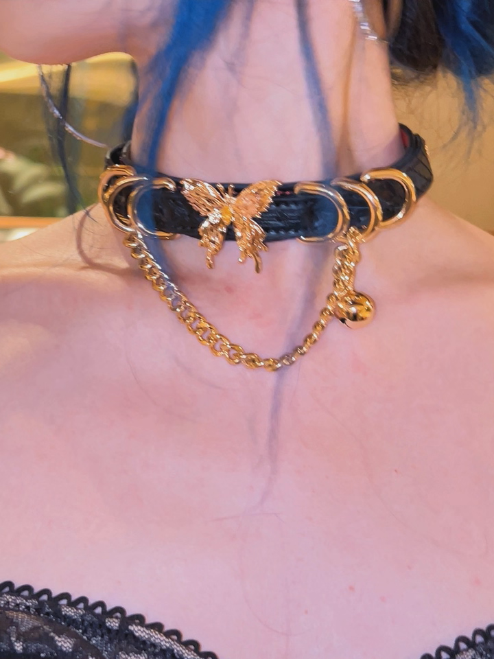 modelled photo of a black, red and gold leather collar featuring an ornate gold butterfly pendant, gold bell, chain and D rings, styled with blue hair and a black lace dress for a glamorous aesthetic.