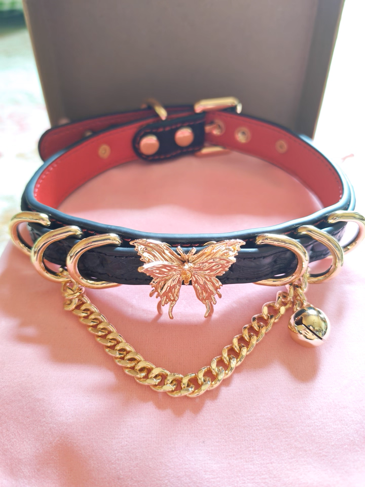 black, red and gold leather collar featuring an ornate gold butterfly pendant, gold bell, D rings and chain, displayed on a pink velvet drawstring bag.