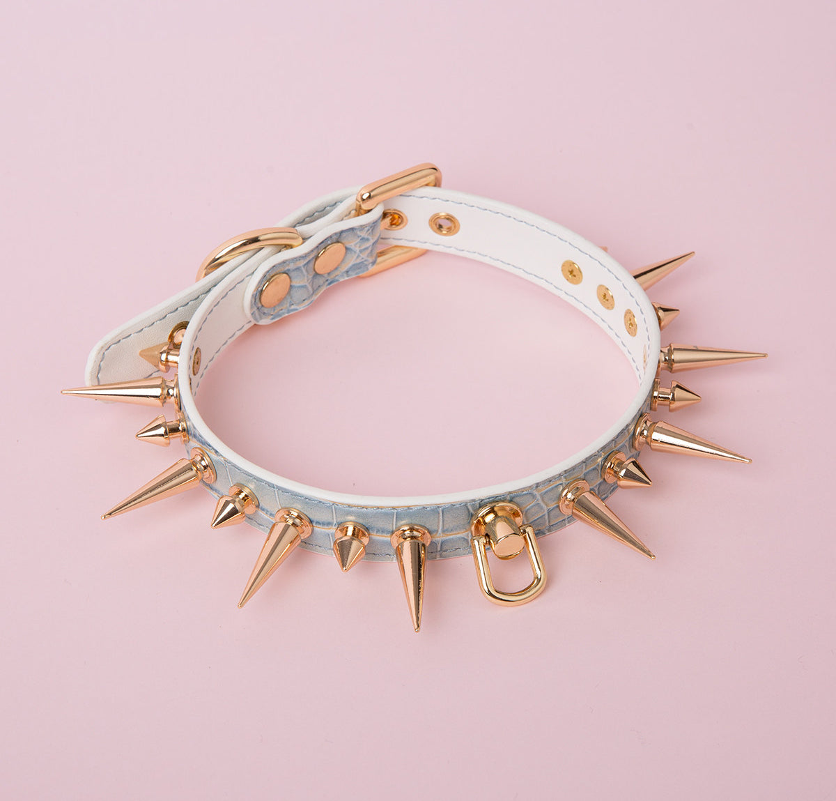 blue and gold leather collar featuring gold-tone alternating long and short spike studs, central D ring attachment and adjustable buckle fastening at the back. displayed against a pink background.