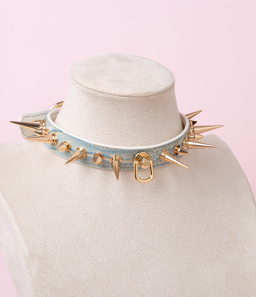 blue and gold leather collar featuring gold-tone alternating long and short spike studs and central D ring attachment. displayed on a white mannequin against a pink background.