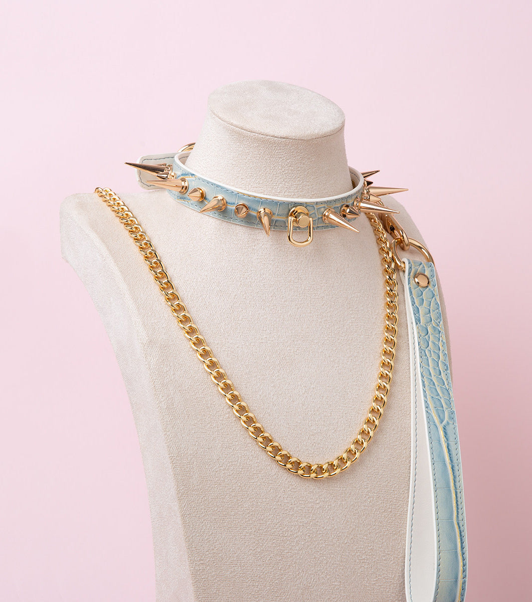 blue and gold leather collar and leash set featuring gold-tone alternating long and short spike studs and D ring attachment with a detachable gold chain leash and blue leather hand strap, displayed on a white mannequin against a pink background.
