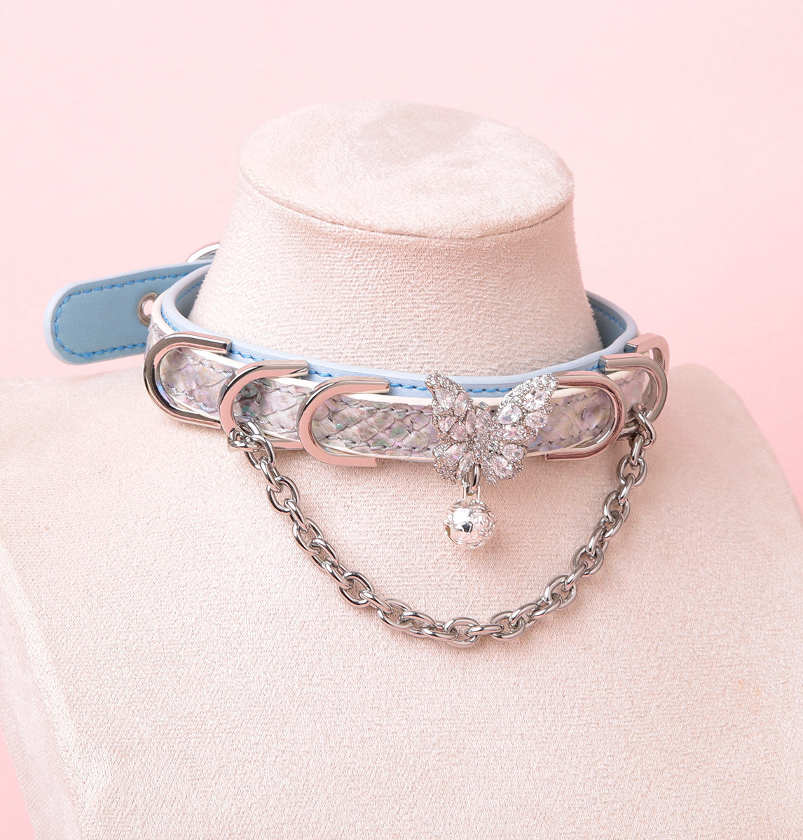 blue and silver leather collar featuring a silver crystal butterfly charm, chains, D rings and bell, displayed on a white mannequin against a pink background.