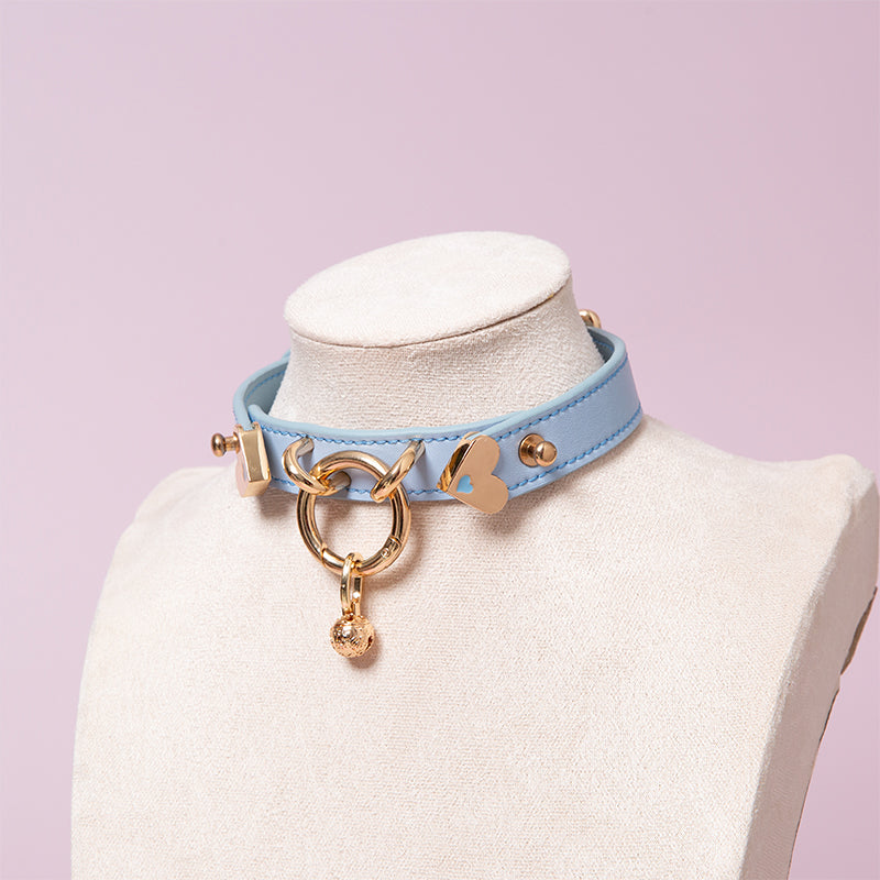 Blue leather collar featuring a central gold tone O ring and detachable gold bell charm. Displayed on a mannequin against a pink background.