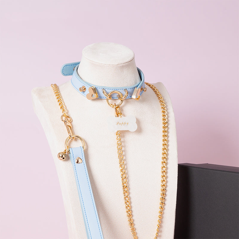 Blue leather collar and leash set featuring a central gold tone O ring, custom engraved bone shaped pet tag, gold chain leash and blue leather hand strap. Displayed on a mannequin against a pink background.