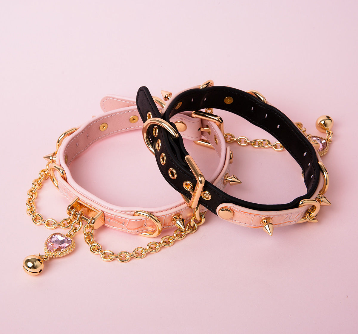 two pink and black leather collars featuring gold and pink crystal heart charms, gold bell, chains and spike studs, displayed on a pink background.