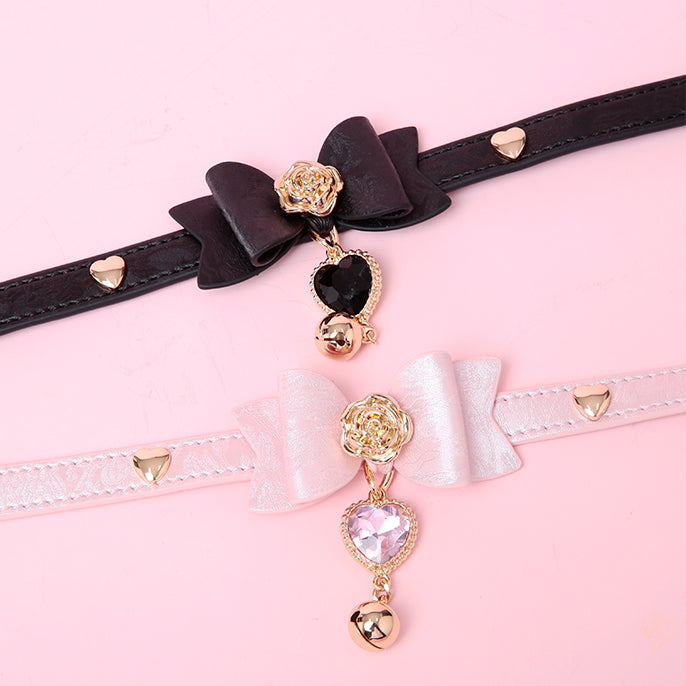 two leather chokers in black and pink, featuring ribbon bow shaped centres, gold rose and heart studs with gold and crystal heart and bell charms, displayed against a pink background.