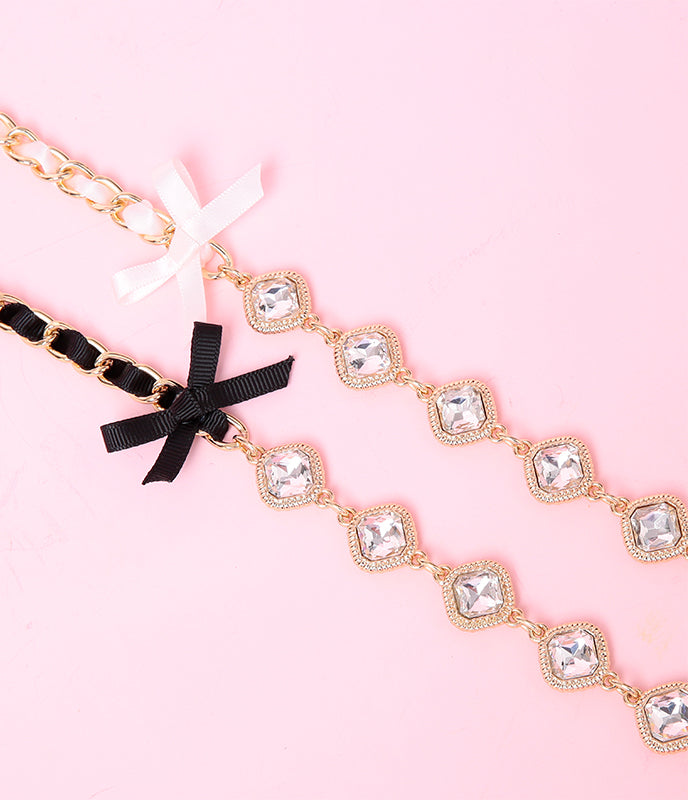close-up of two detachable chain and crystal leashes in pink and black, featuring ribbon threaded through the chain, satin bow applique and gold and clear crystal charms, displayed against a pink background.