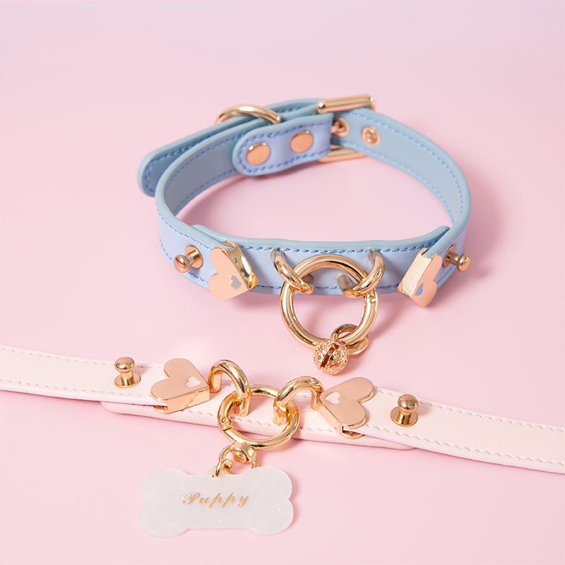 Two blue and pink leather collars featuring a central gold tone O ring, heart shaped hardware, detachable custom engraved bone shaped pet tag and gold bell charm. Displayed on a pink background.