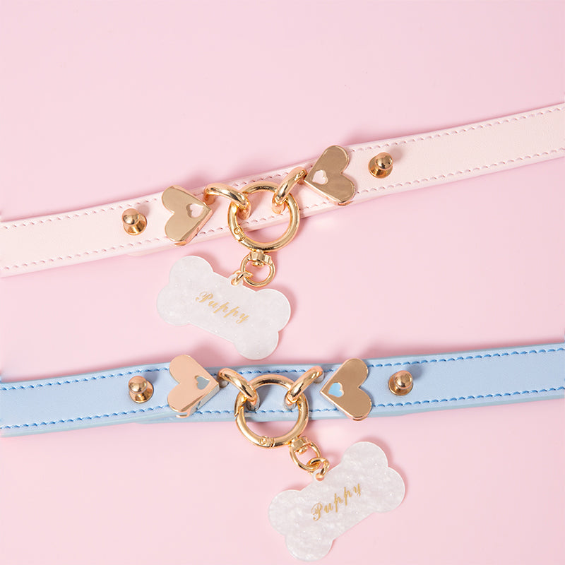 Two blue and pink leather collars featuring a central gold tone O ring, heart shaped hardware and detachable custom engraved bone shaped pet tag. Displayed on a pink background.